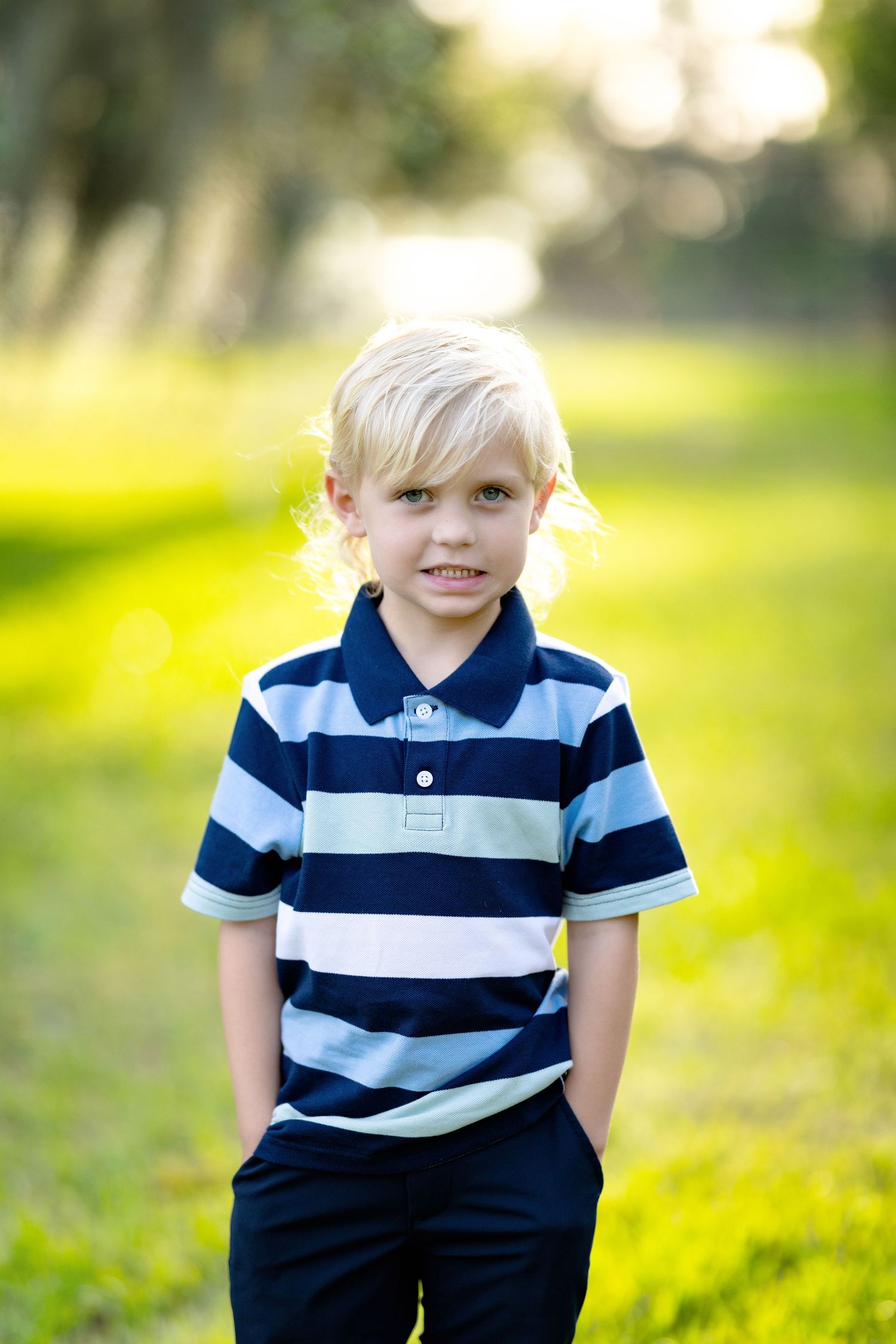 Smiling blond boy in striped navy polo shirt standing outdoors in a sunny green park with hands in pockets.