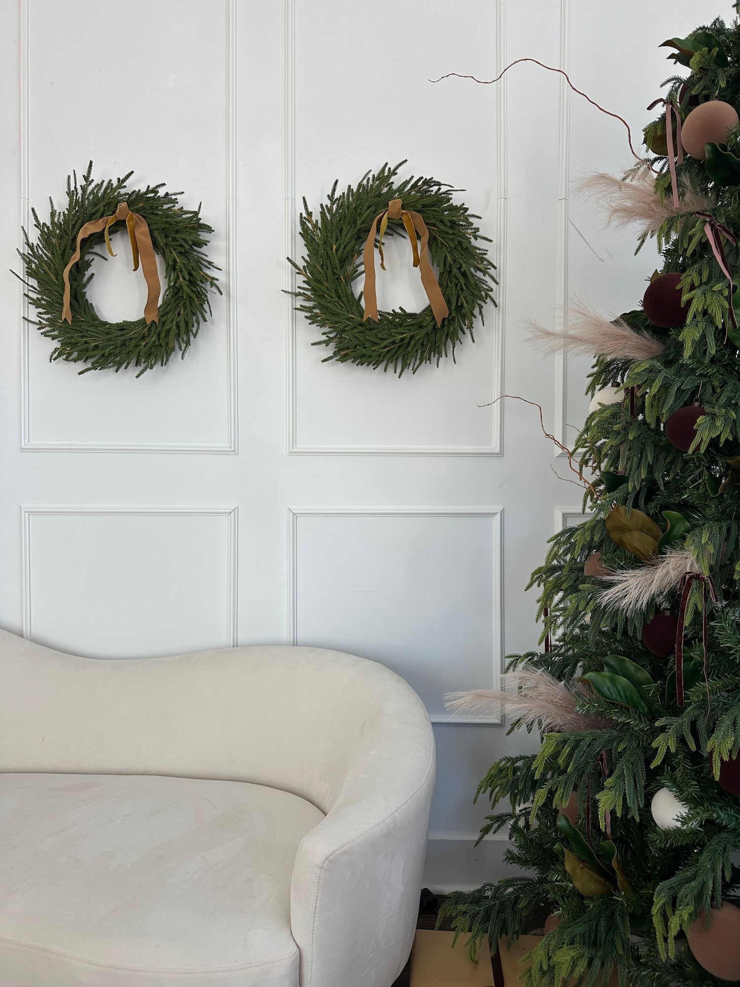Elegant holiday decor with two green wreaths on white wall, cream sofa, and decorated Christmas tree with feathers.