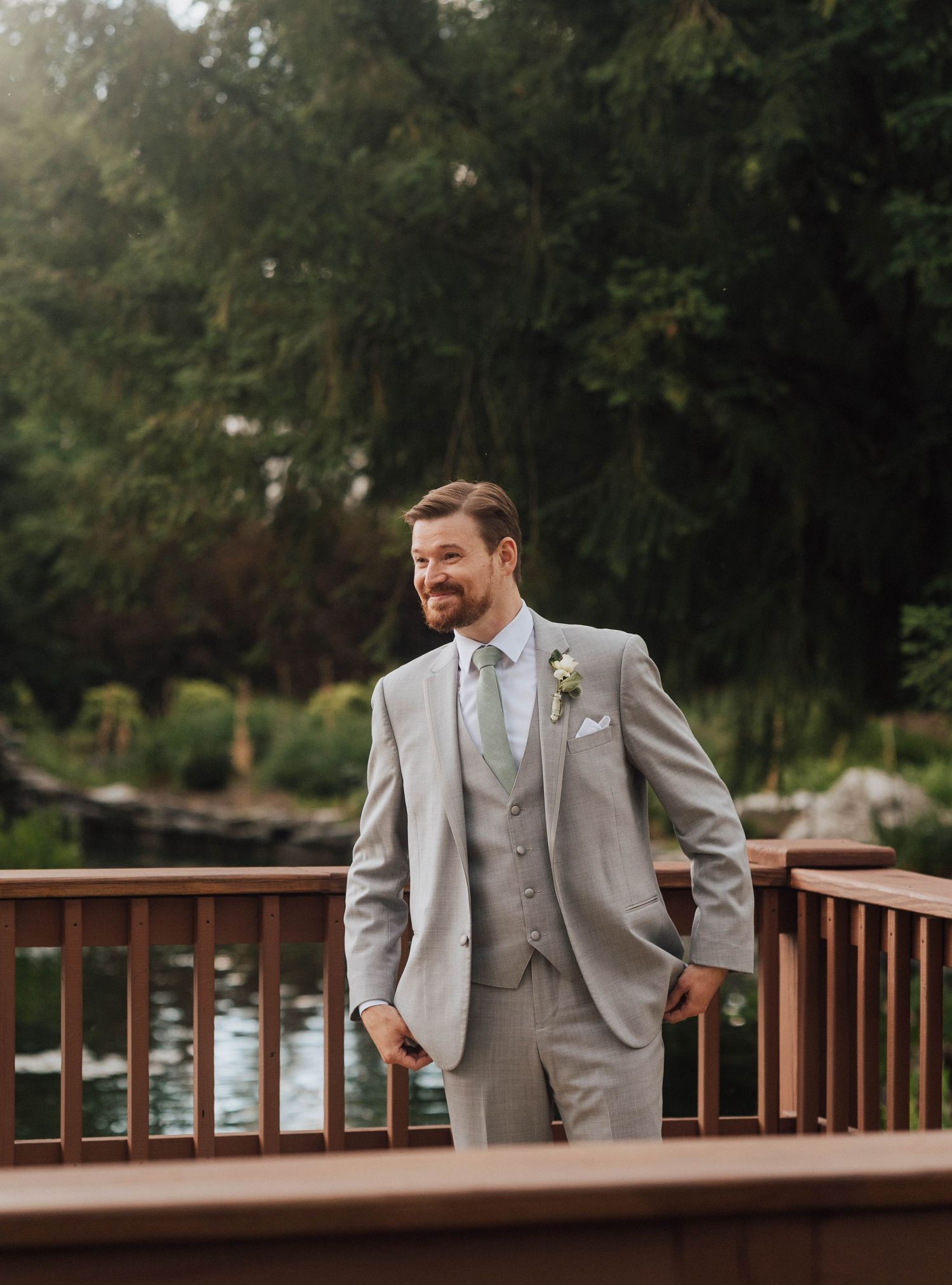 Smiling groom in light gray three-piece suit with boutonniere standing on wooden deck surrounded by lush greenery.