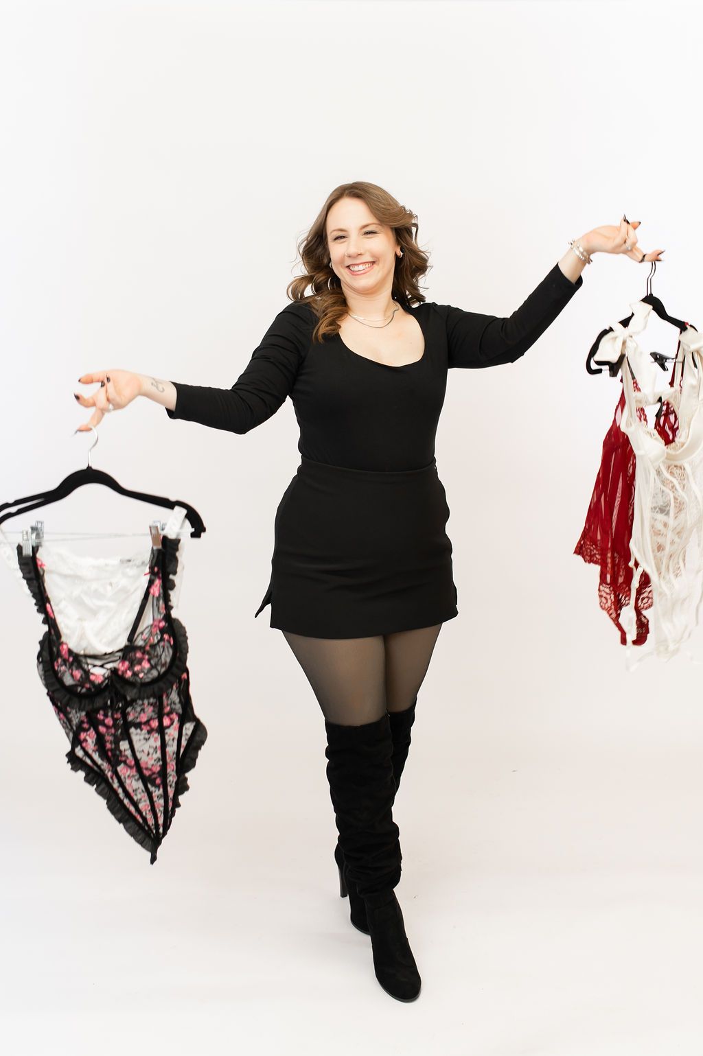 Smiling woman in black dress and boots holding up lingerie on hangers against white background.
