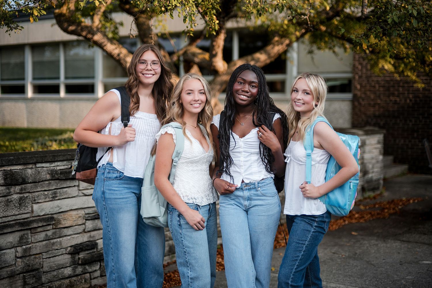 Four smiling teenage girls with backpacks pose together outdoors on a sunny day near a school building.