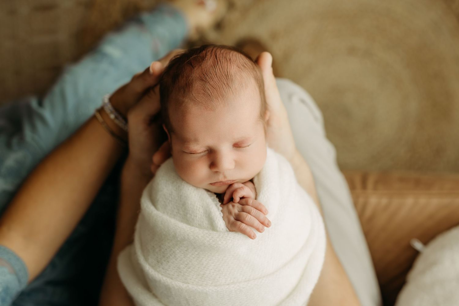 Newborn baby swaddled in white wrap sleeping peacefully while mother gently cradles head in warm natural light.