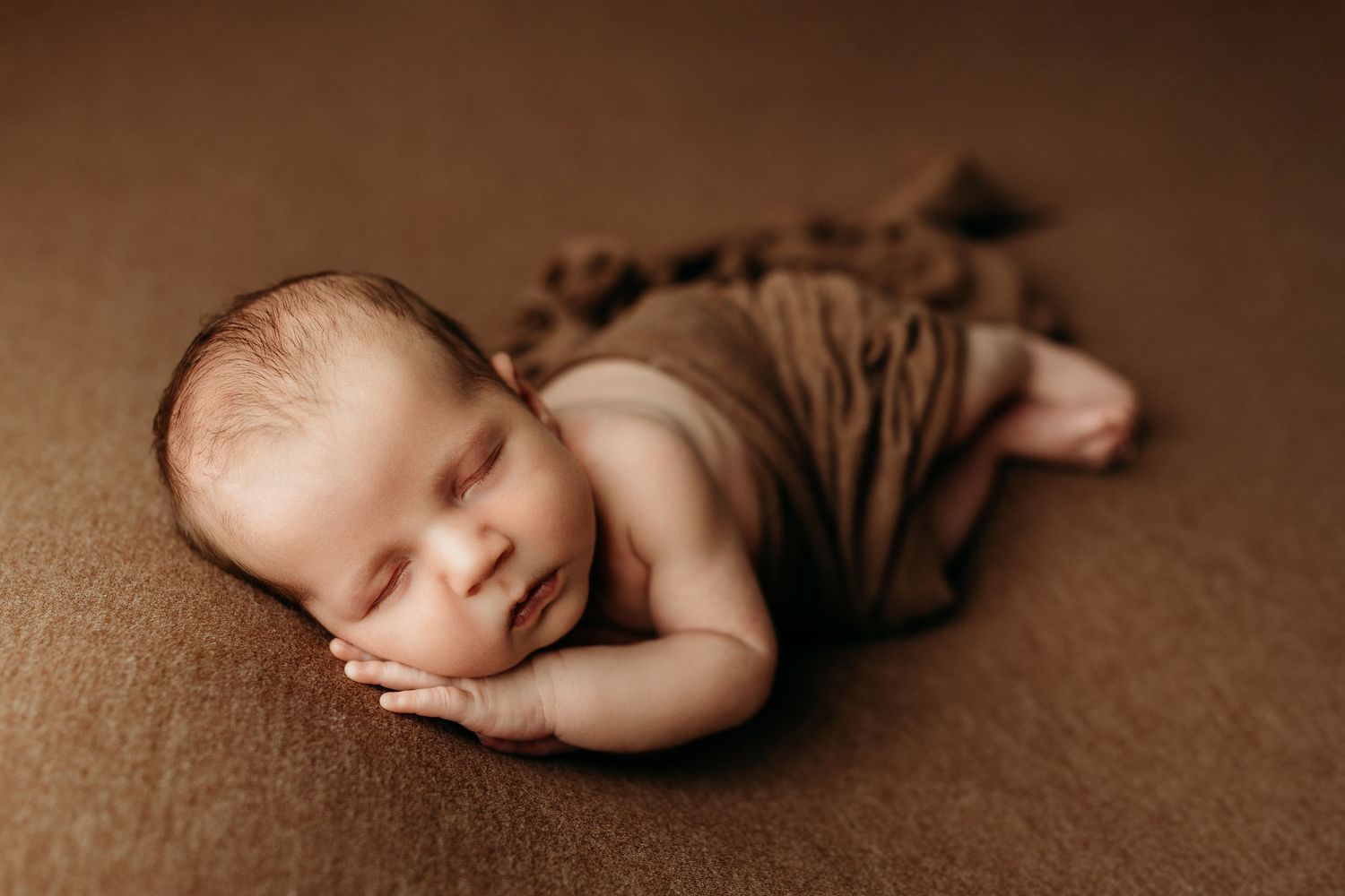 Peaceful newborn baby wrapped in brown swaddle sleeping on brown backdrop in professional studio portrait.
