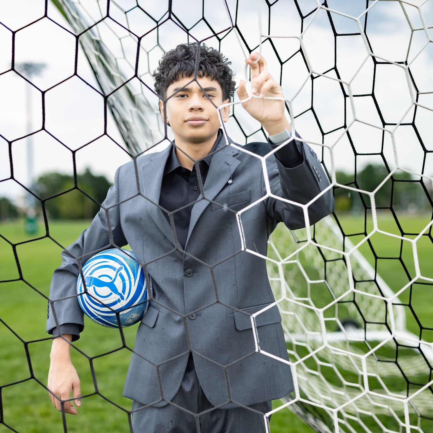 Young man in gray suit holding soccer ball poses confidently in front of a soccer goal net outdoors.