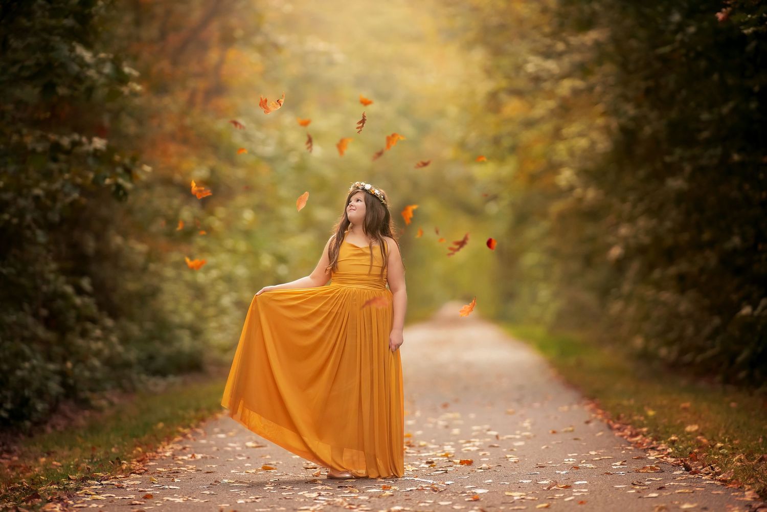 Woman in a flowing mustard-yellow maxi dress stands on a tree-lined path as golden autumn leaves drift through the warm, glowing air.