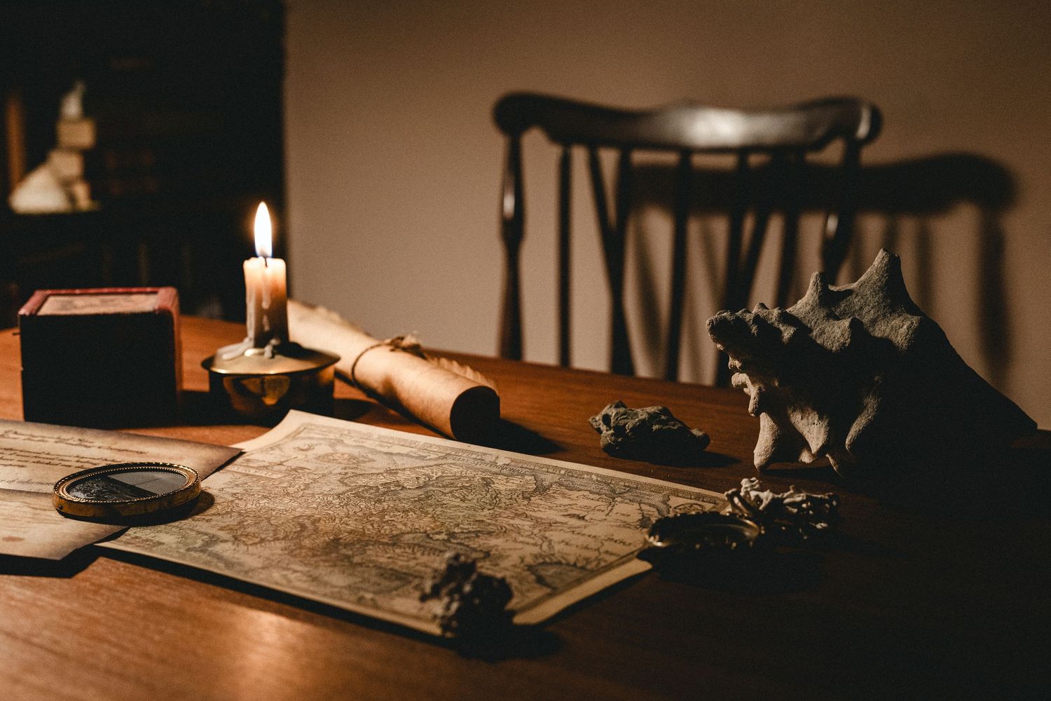 Antique map on wooden desk with lit candle, magnifying glass, scroll, and decorative skull figurine in dim light.