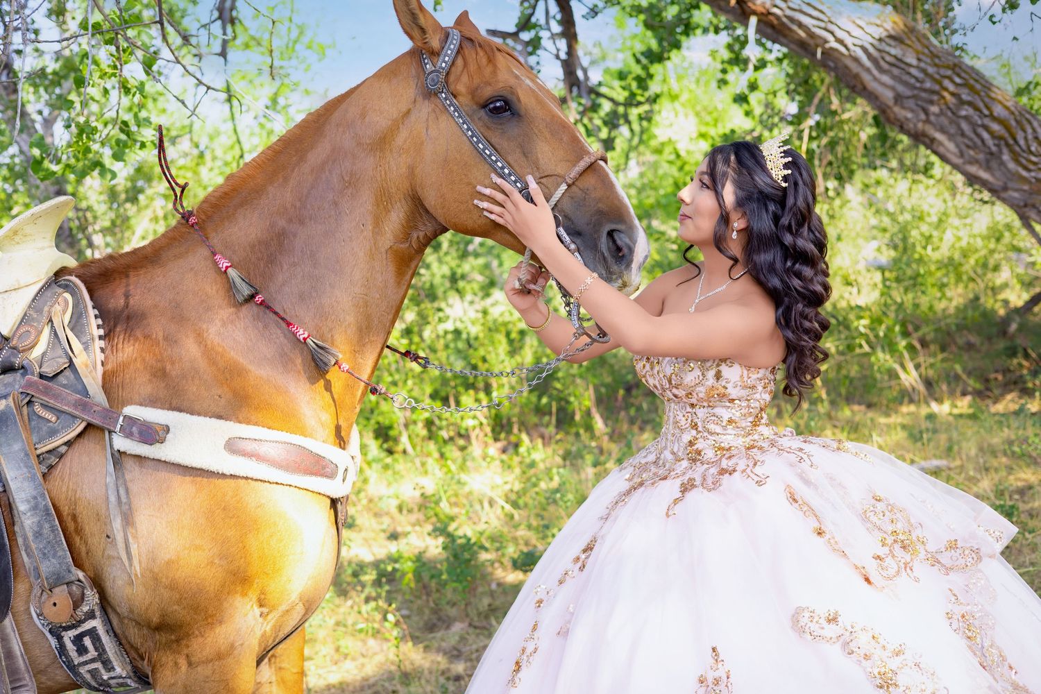 A young woman in a white and gold quinceañera gown gently touches a chestnut horse outdoors.