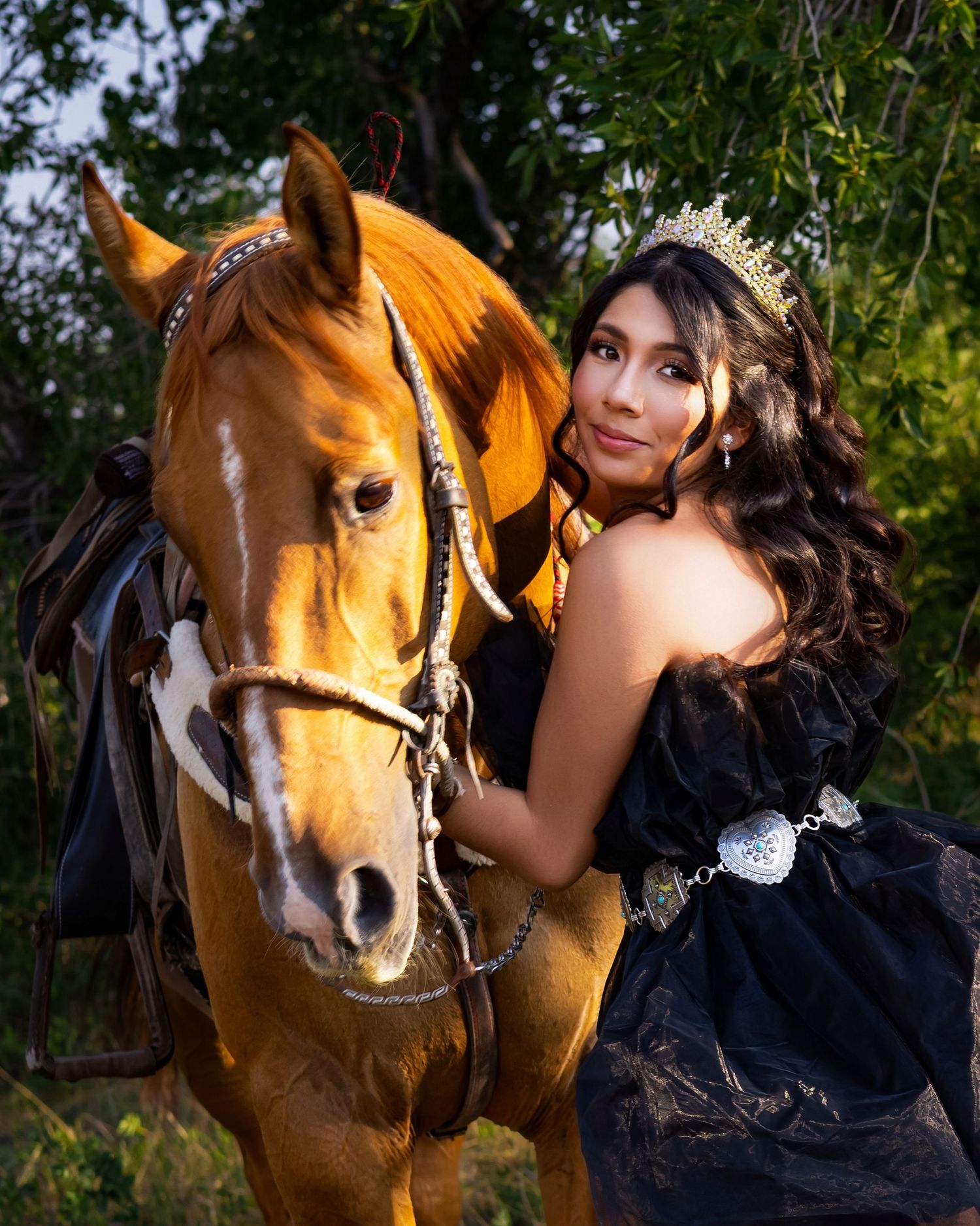 Young woman in black gown and tiara poses elegantly with a chestnut horse outdoors among green trees.