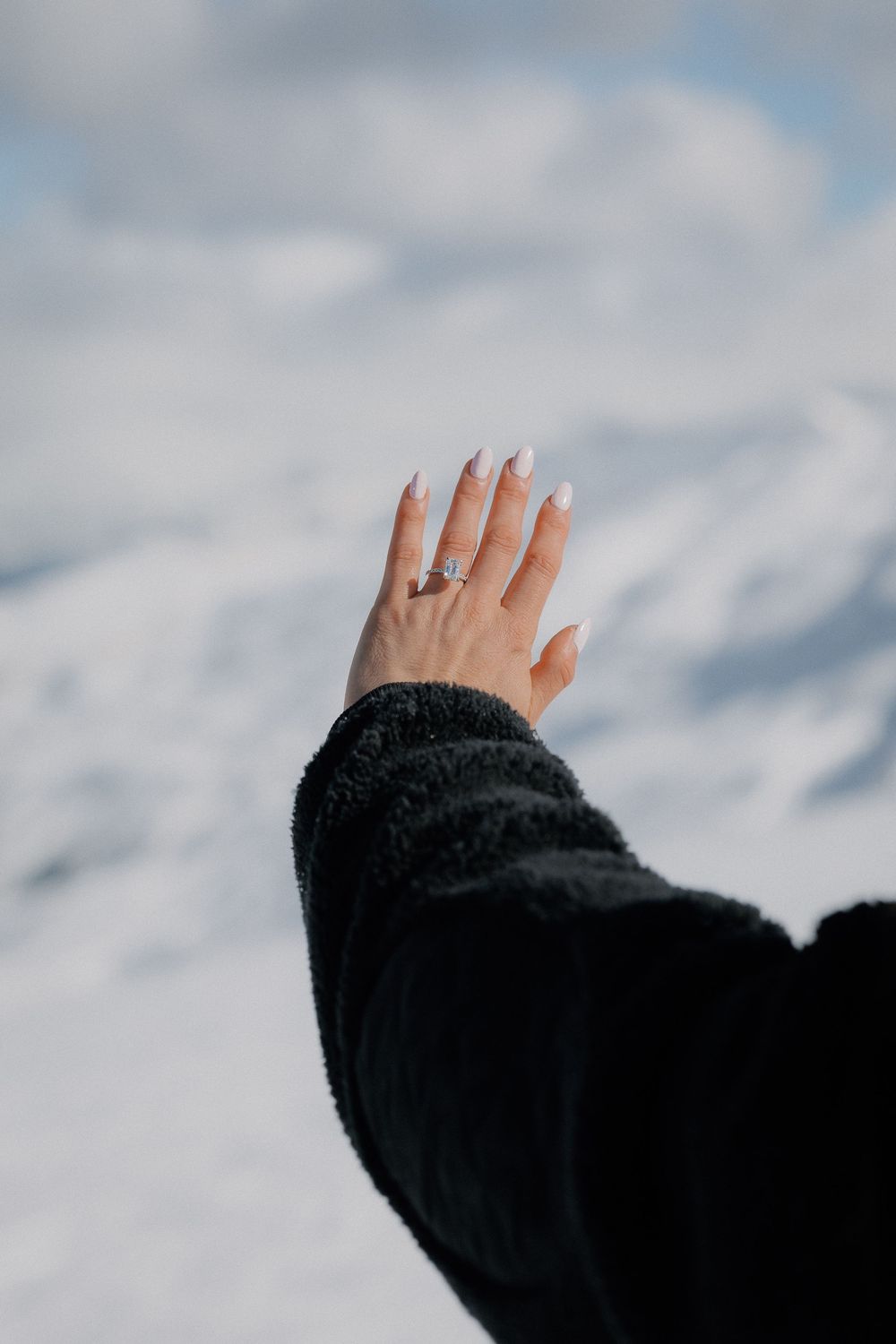 A hand with white manicured nails and a diamond ring raised against a snowy mountain backdrop, wearing a black fur sleeve.