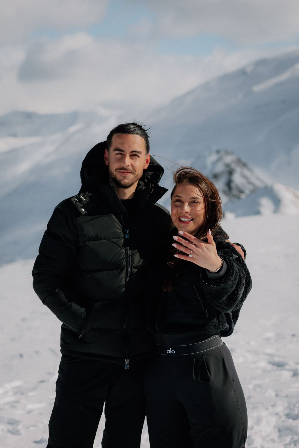 Couple in black jackets showing engagement ring on snowy mountain backdrop, smiling at camera.