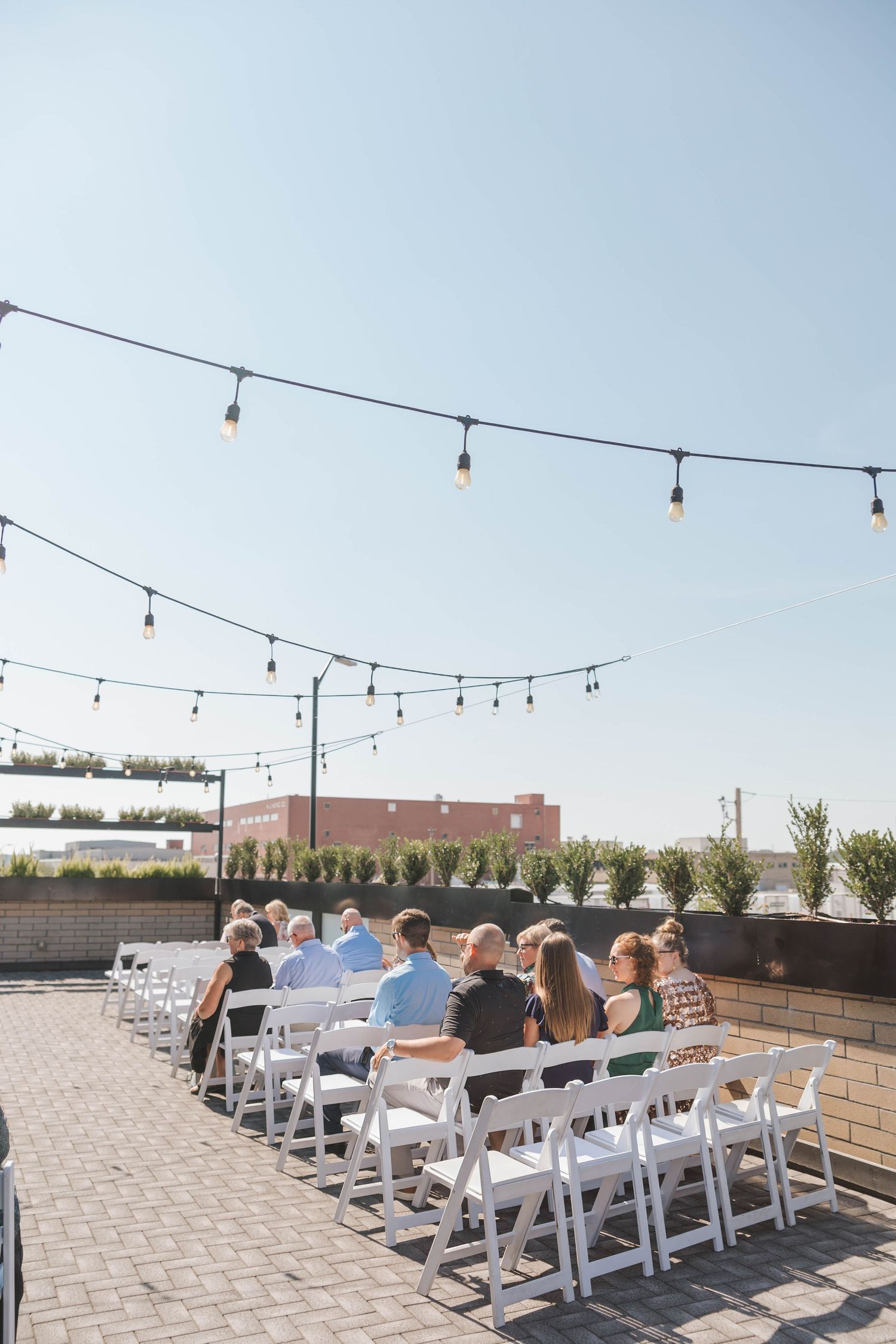 Bride in white gown walks down outdoor aisle with flower girl under string lights at sunny wedding ceremony.