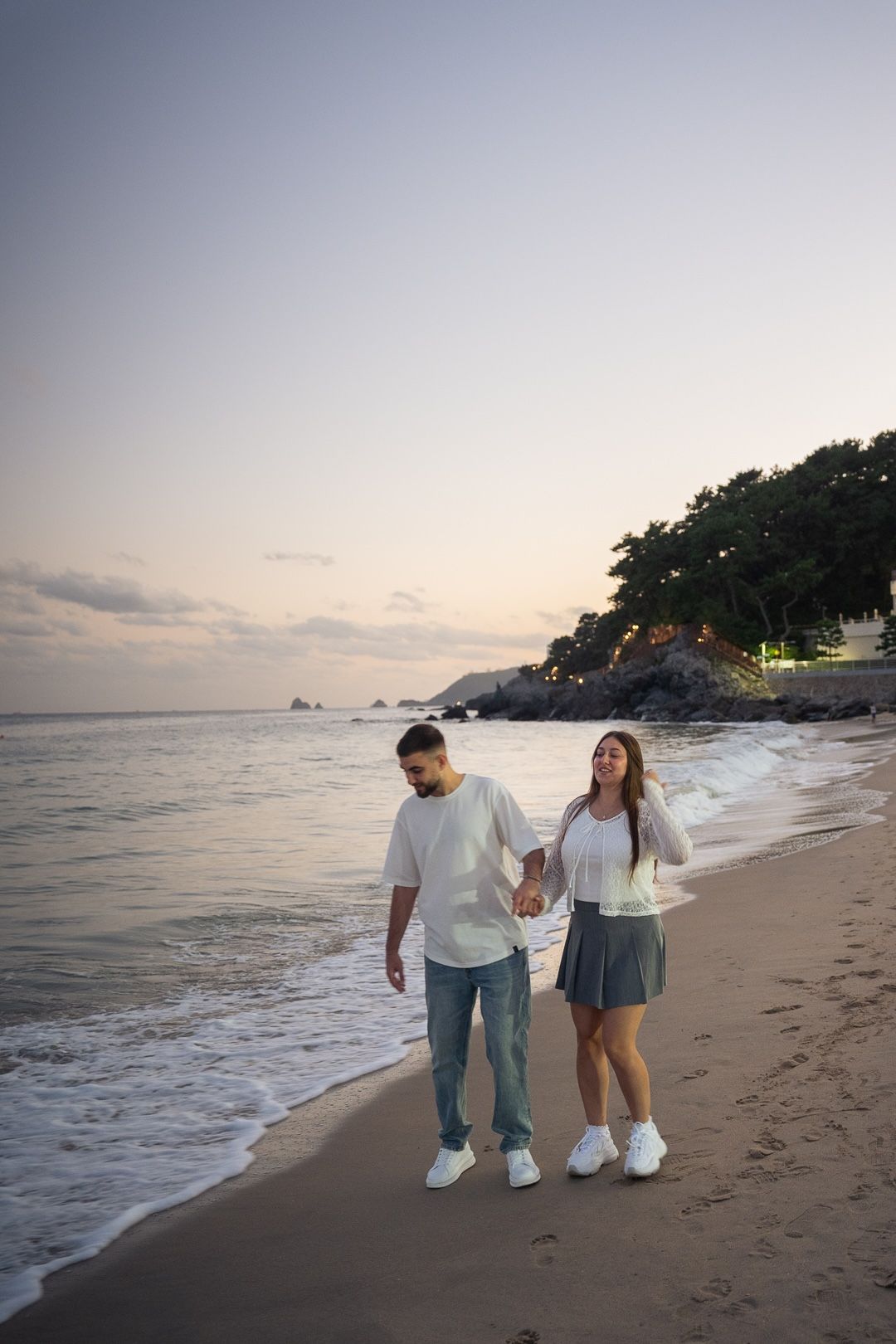 Couple holding hands walking on a sandy beach at sunset with ocean waves and a rocky tree-lined shore in the background.