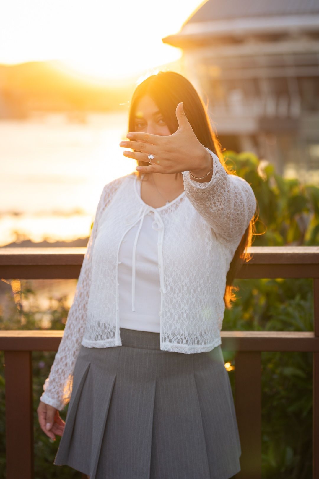 Woman in white lace cardigan and gray skirt poses at sunset, shielding camera with outstretched hand outdoors.