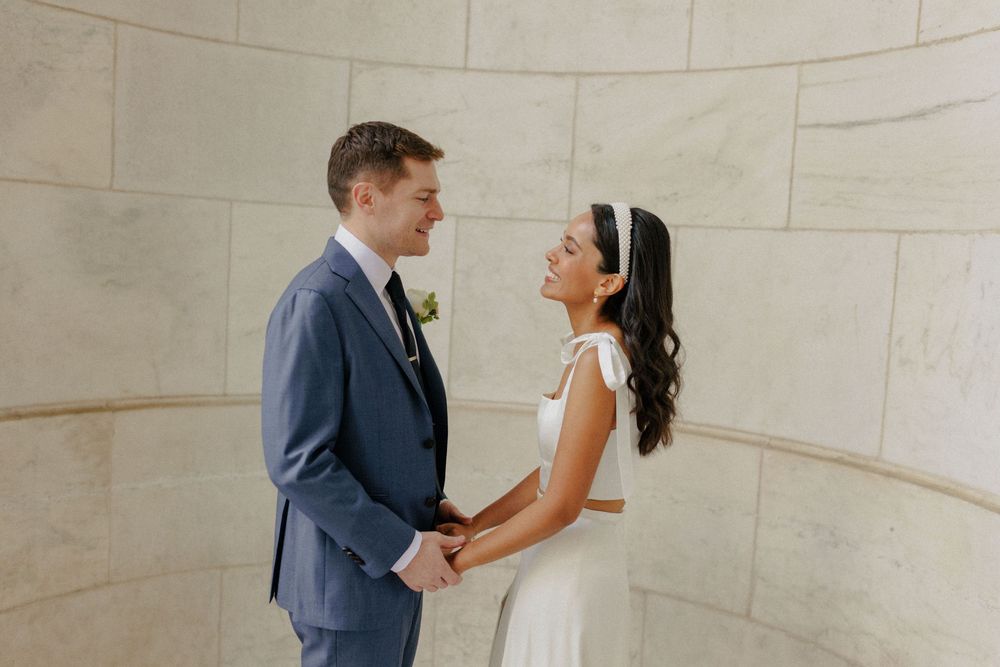 A bride and groom hold hands and smile at each other in a bright marble venue on their wedding day.