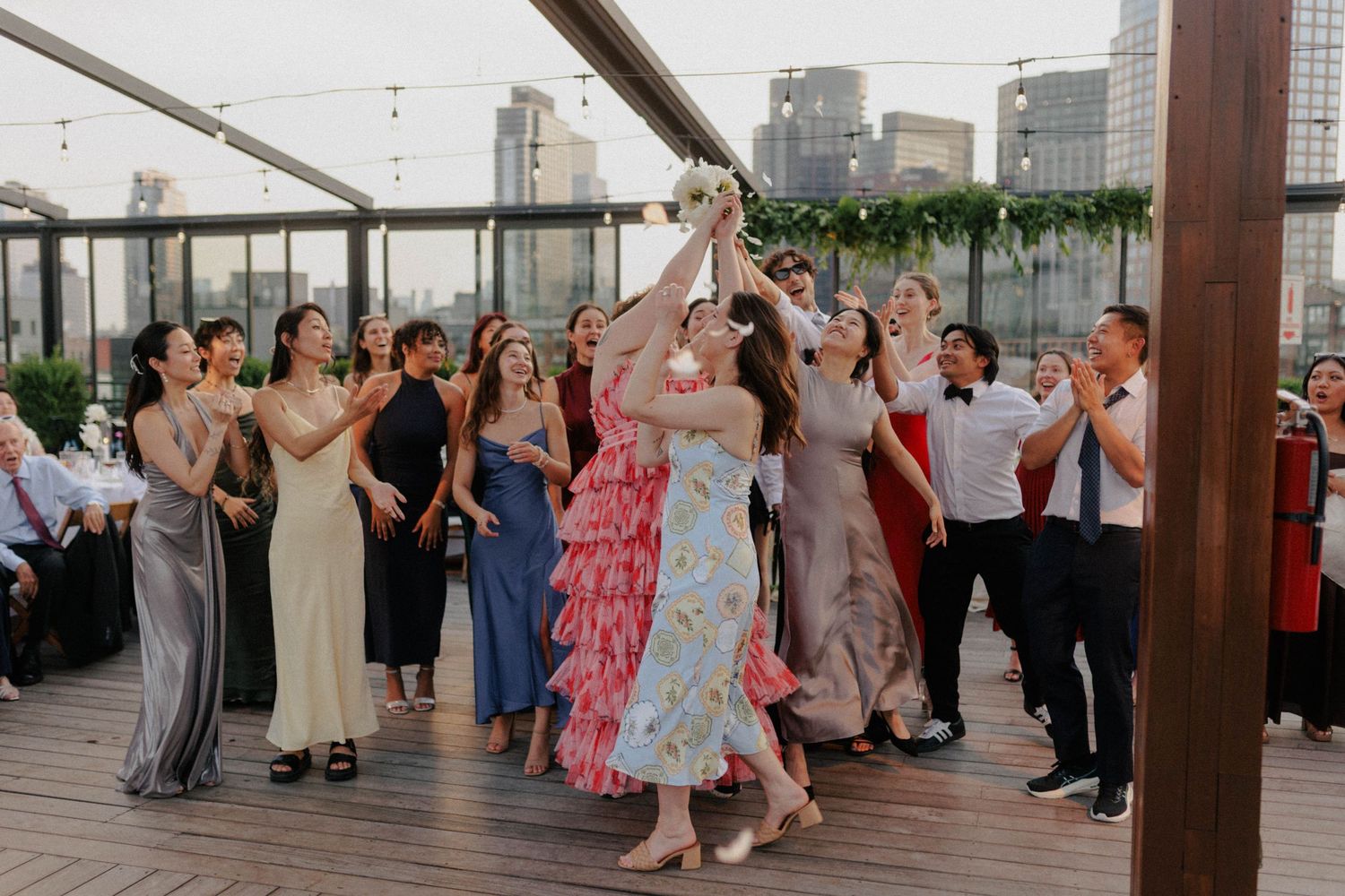 Guests reach for the bouquet toss at a rooftop wedding reception with city skyline views.