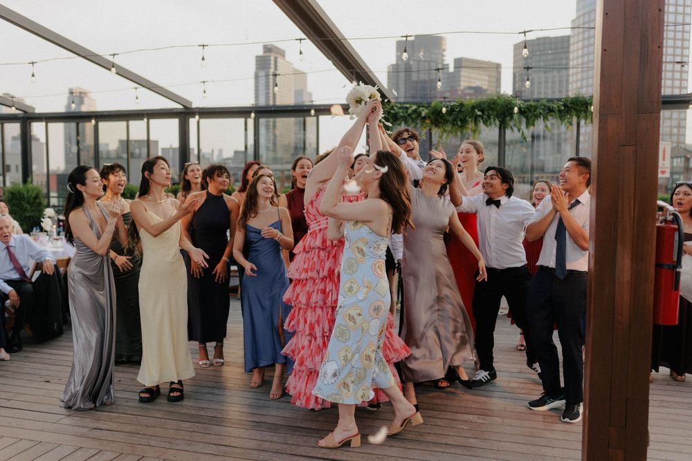 Guests reach for the bouquet toss at a rooftop wedding reception with city skyline views.