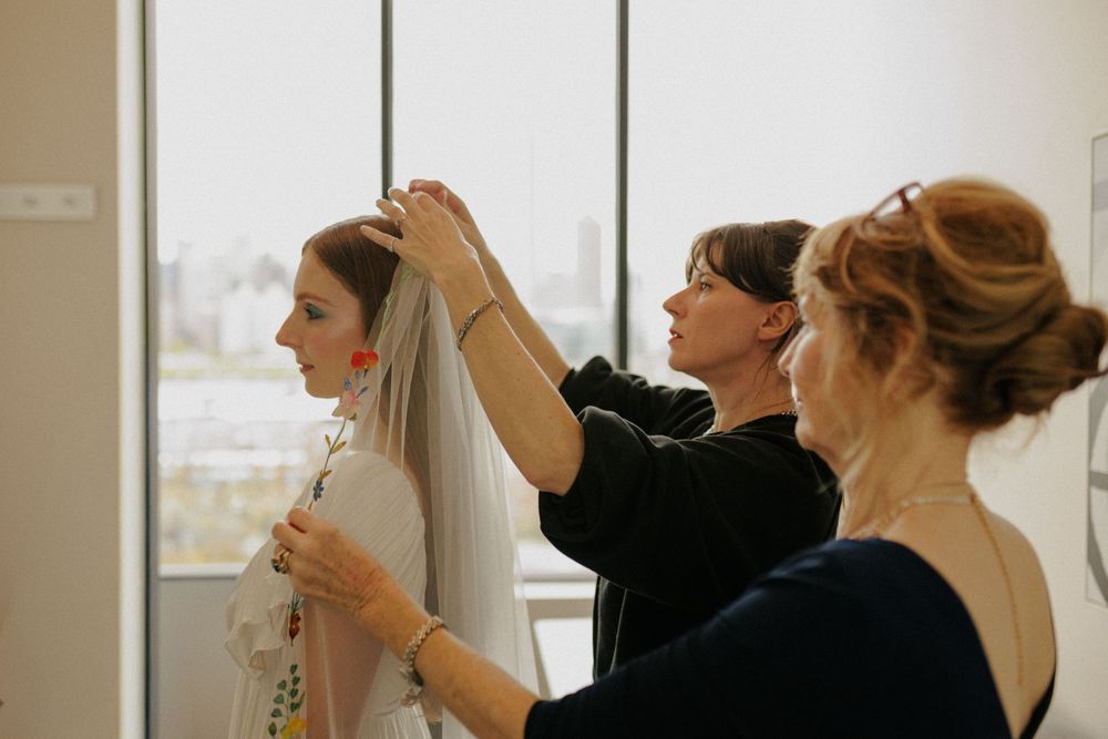 Two women help a bride put on her wedding veil near a large window with a city view.
