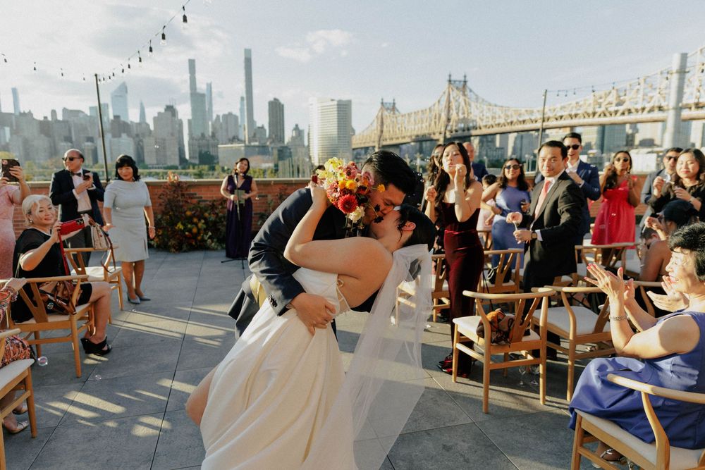Bride and groom share first kiss at rooftop wedding ceremony with NYC skyline and Queensboro Bridge backdrop.