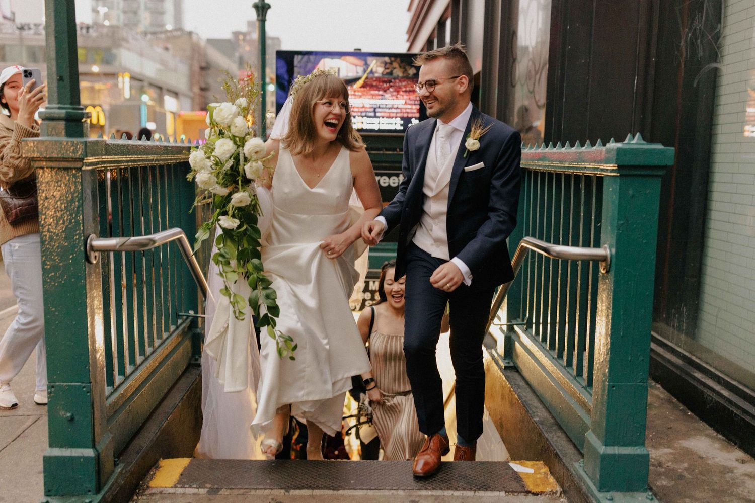Bride and groom laughing while descending NYC subway stairs in wedding attire, adorned with white floral bouquet.