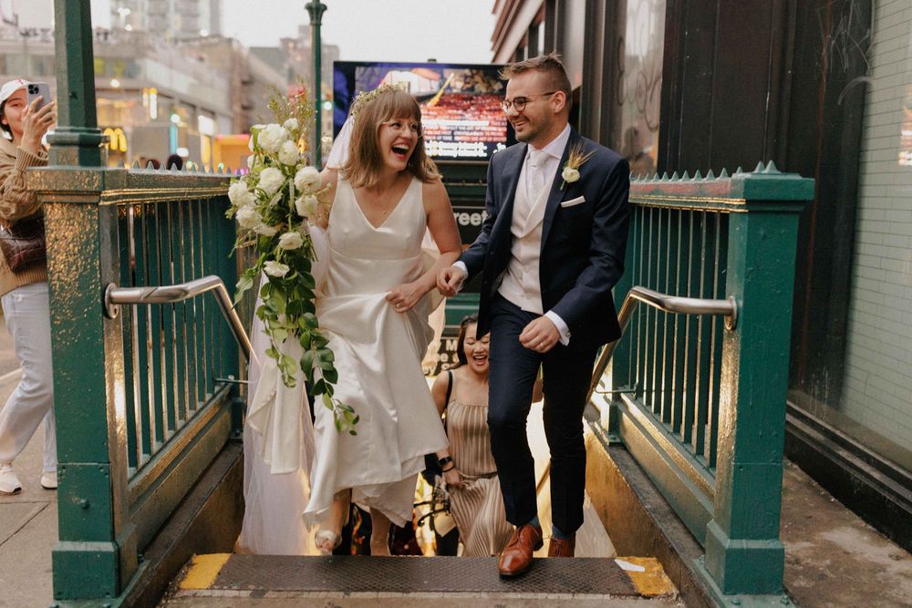 Bride and groom laughing while descending NYC subway stairs in wedding attire, adorned with white floral bouquet.