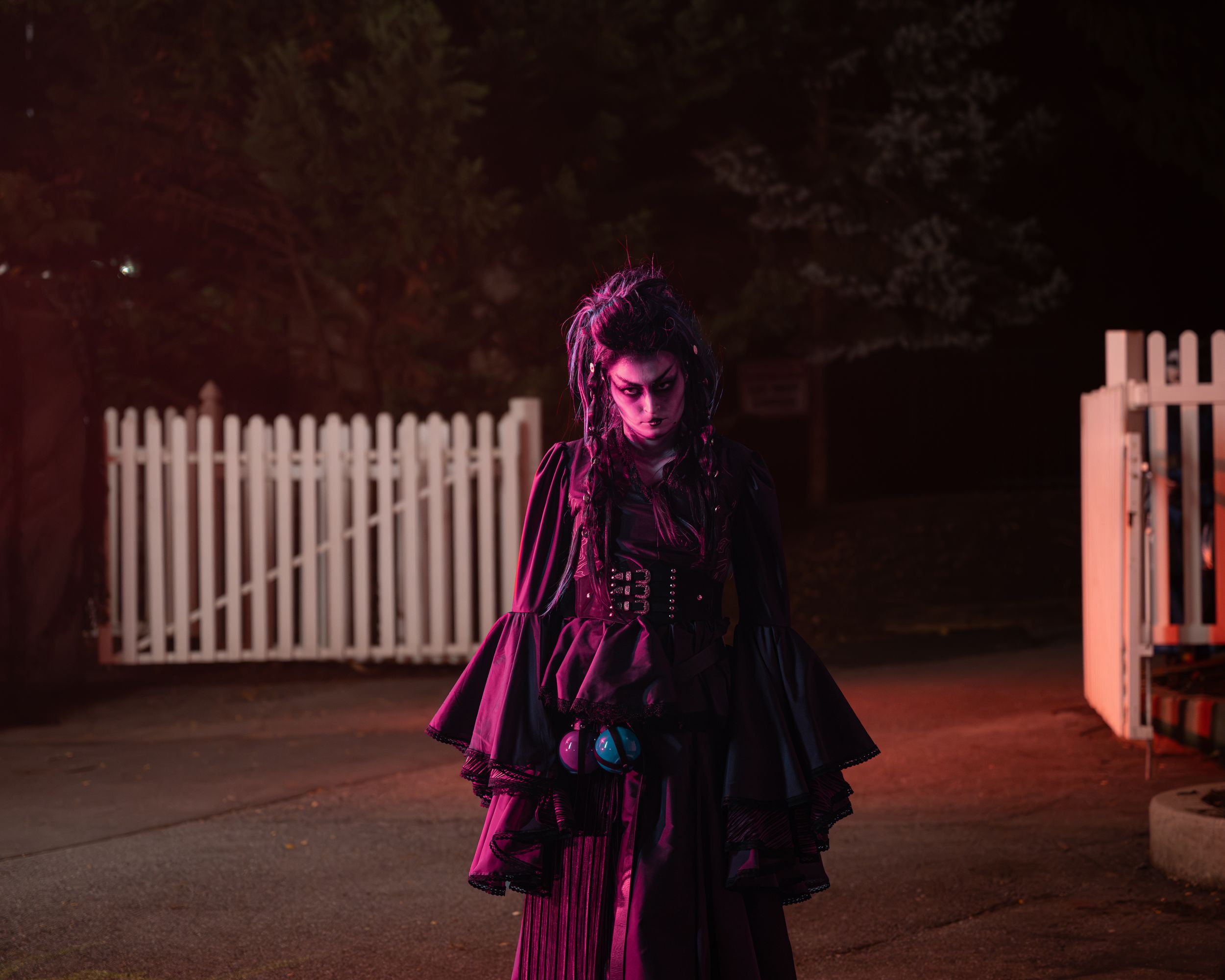 Person in gothic Halloween costume with dark cape and corset stands near white picket fence at night.