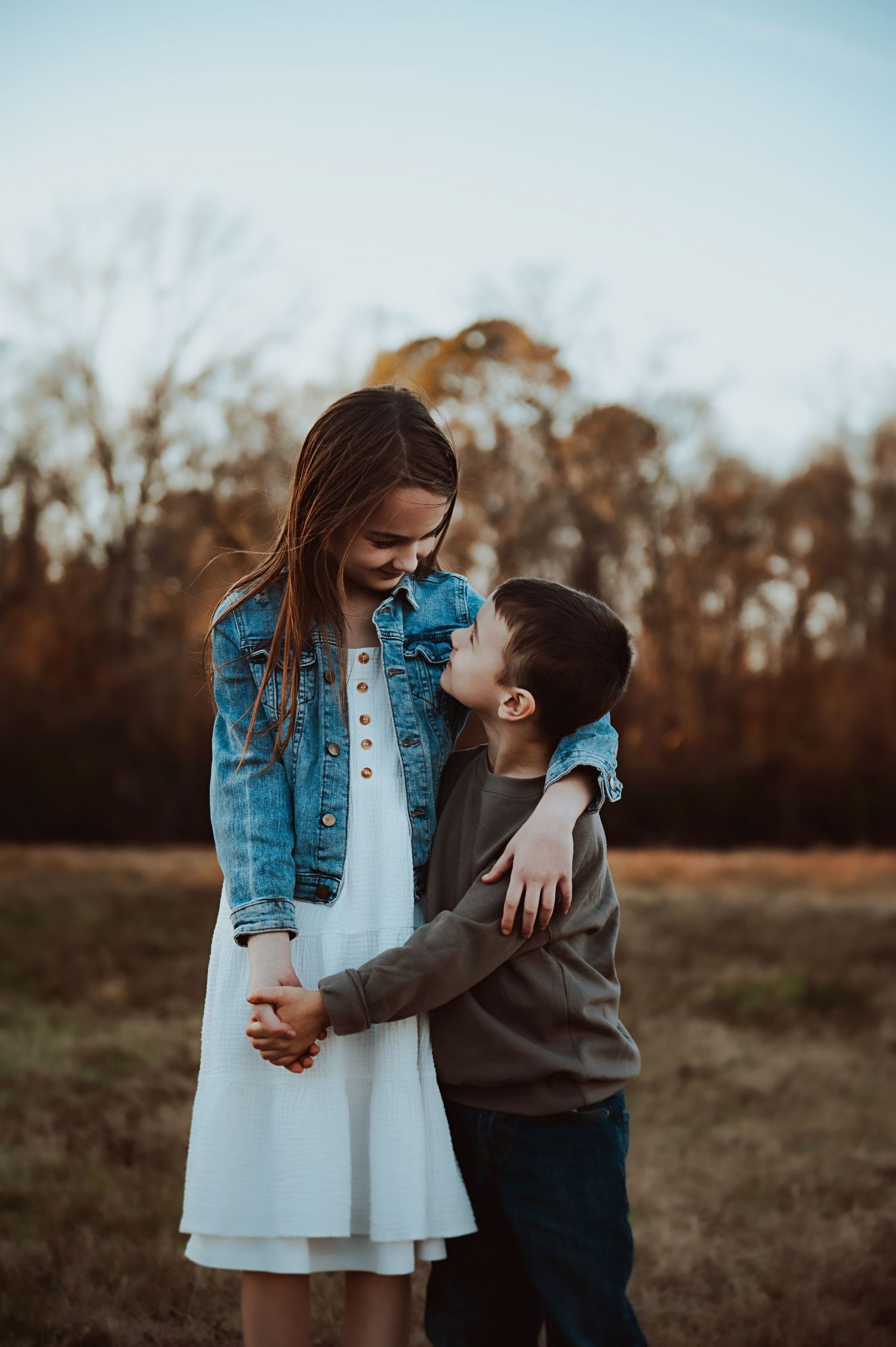 Girl in a denim jacket and white dress hugs a smiling little boy in a gray sweater in a grassy field with autumn trees.