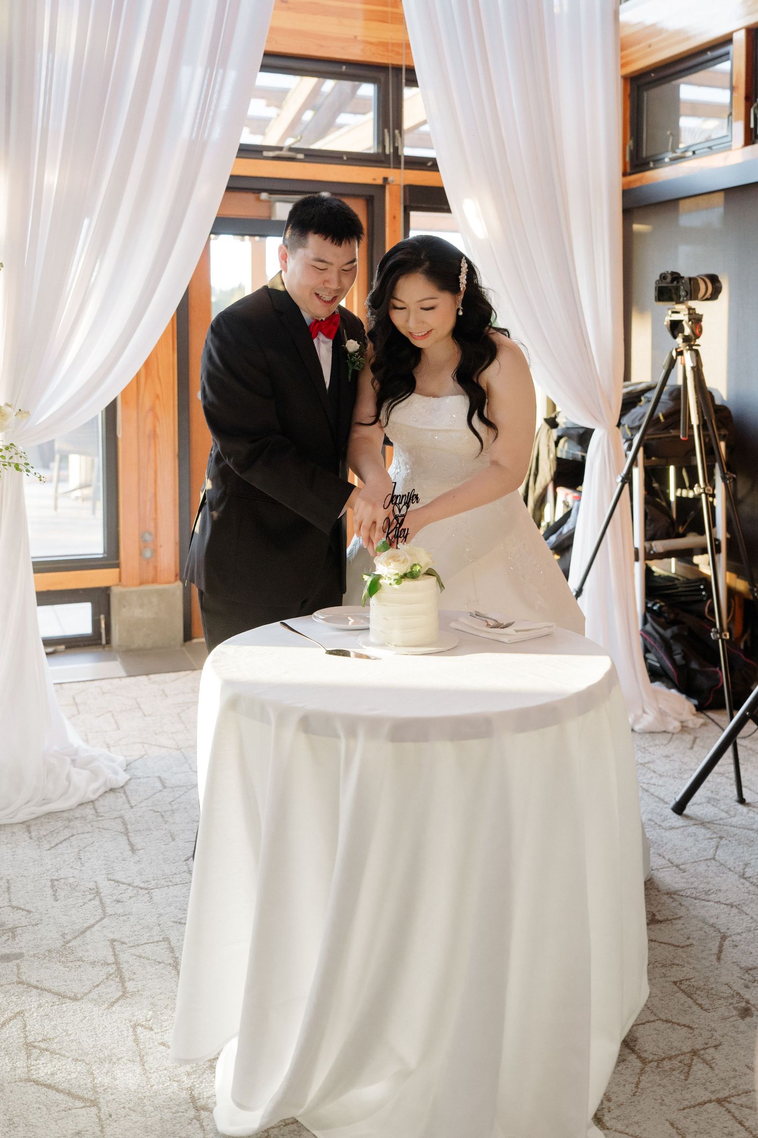 Bride in a white gown and groom in a black suit with red bow tie cut their wedding cake together at a draped table.
