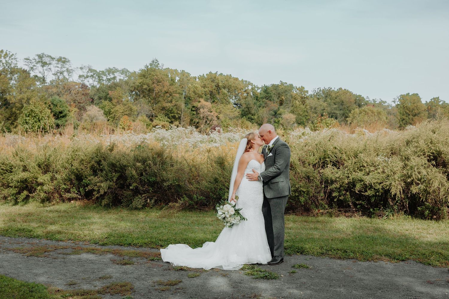 Bride and groom share a romantic embrace in a lush outdoor setting with golden meadows and trees on their wedding day.