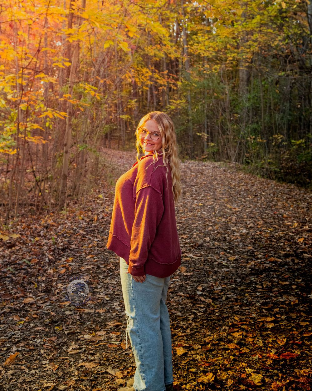 Woman with long blonde hair in a burgundy sweater and jeans smiles over her shoulder on a leaf-covered forest path in golden autumn light.