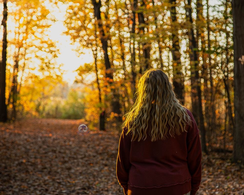 Woman with long wavy hair in a maroon sweater stands on a leaf-covered forest path surrounded by golden autumn trees.