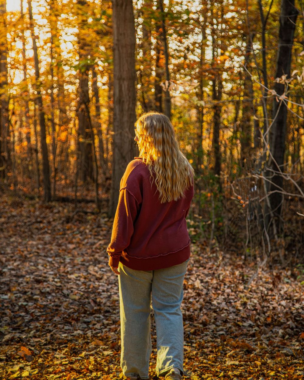 Woman with long blonde hair in a maroon sweater walks through a sunlit autumn forest covered in fallen leaves.