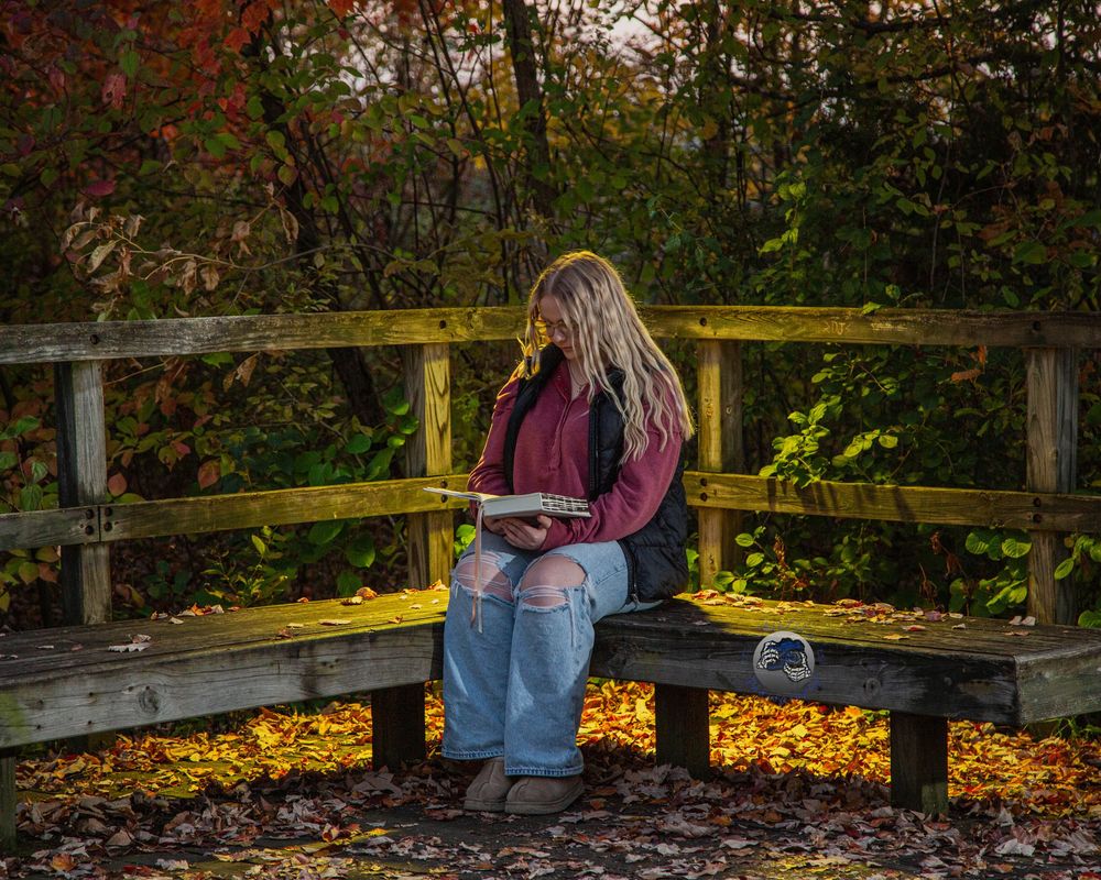 Woman with long blonde hair reading a book on a wooden park bench surrounded by colorful autumn leaves and trees.