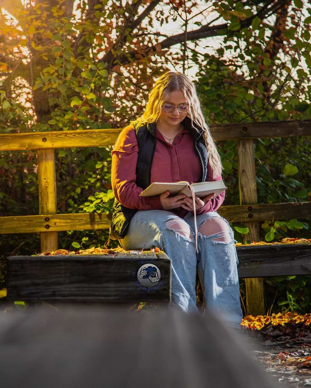 Young woman with long blonde hair and glasses sits on a wooden bench reading a book outdoors surrounded by autumn leaves.
