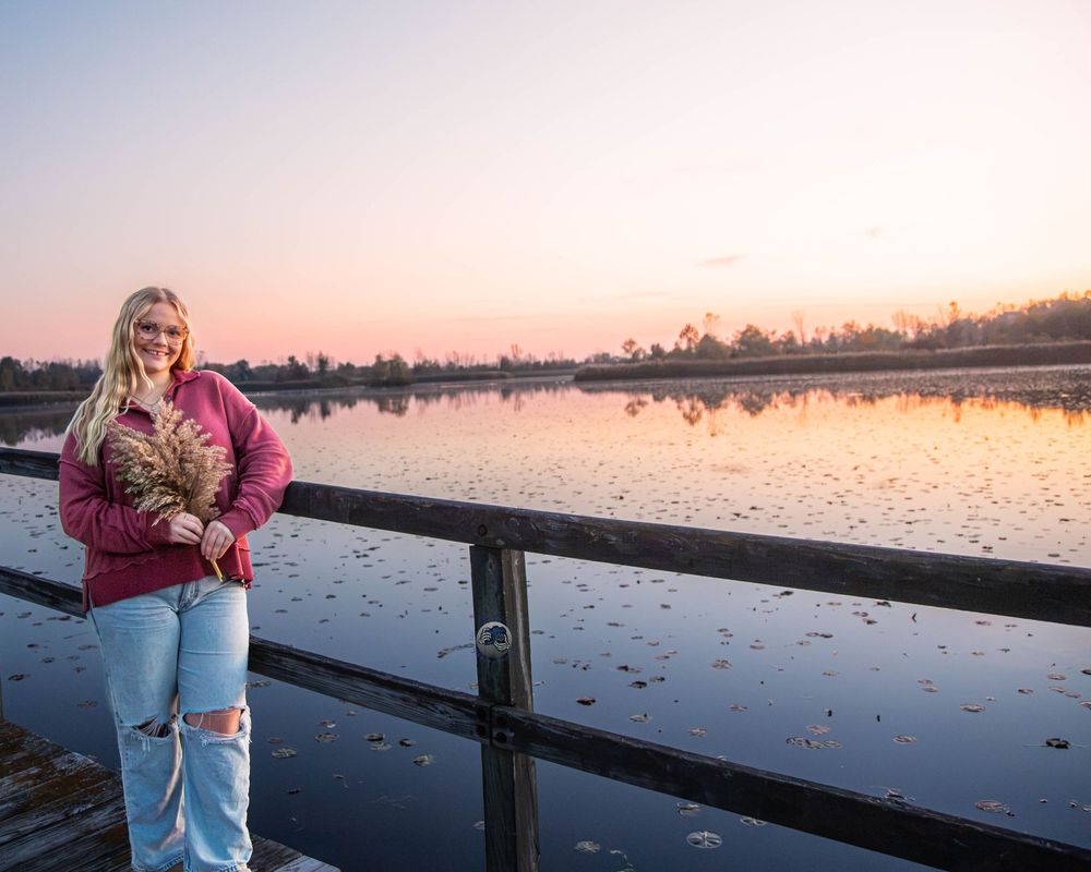 Young woman in a maroon sweater and ripped jeans holds dried grasses on a wooden dock overlooking a calm lake at sunset.
