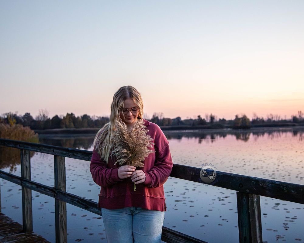 Woman in a burgundy sweater stands on a wooden lakeside dock holding pampas grass, with a soft pink sunset sky reflected on the water.