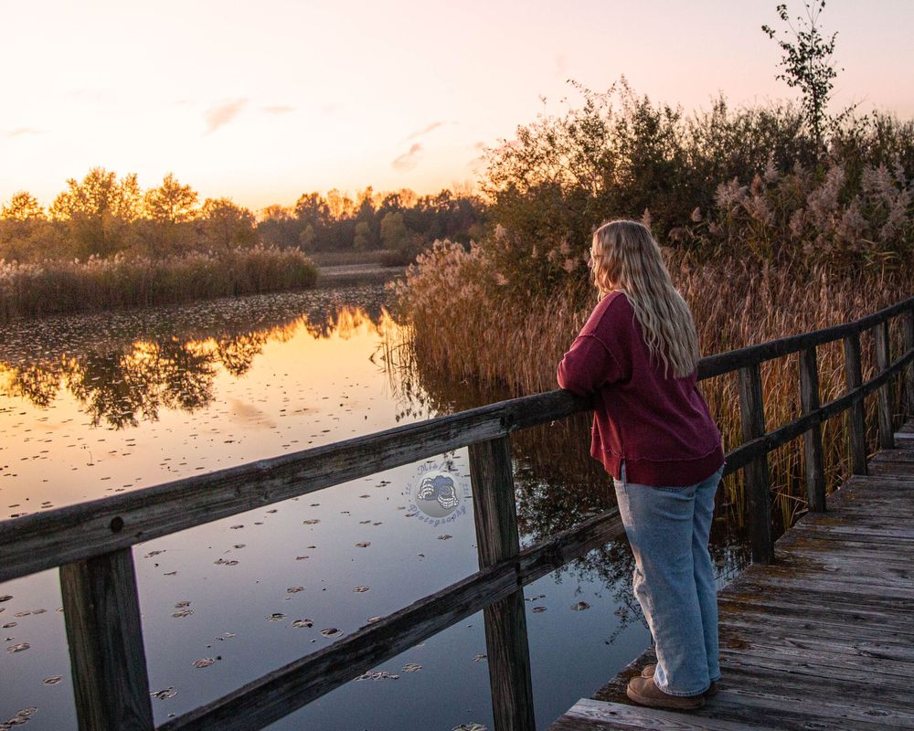 Woman in a maroon sweater leans on a wooden railing, gazing over a still pond and golden marsh grasses at sunset.