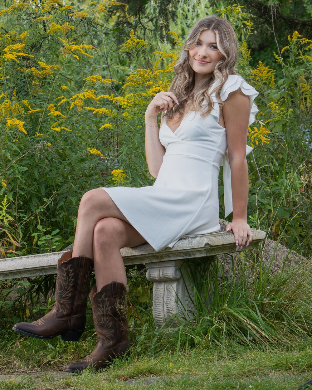 Young woman in white dress and brown cowboy boots sits on a stone bench surrounded by lush green and yellow garden foliage.