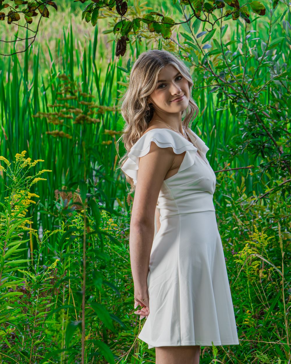 Young woman in white ruffle dress posing among lush green plants outdoors.