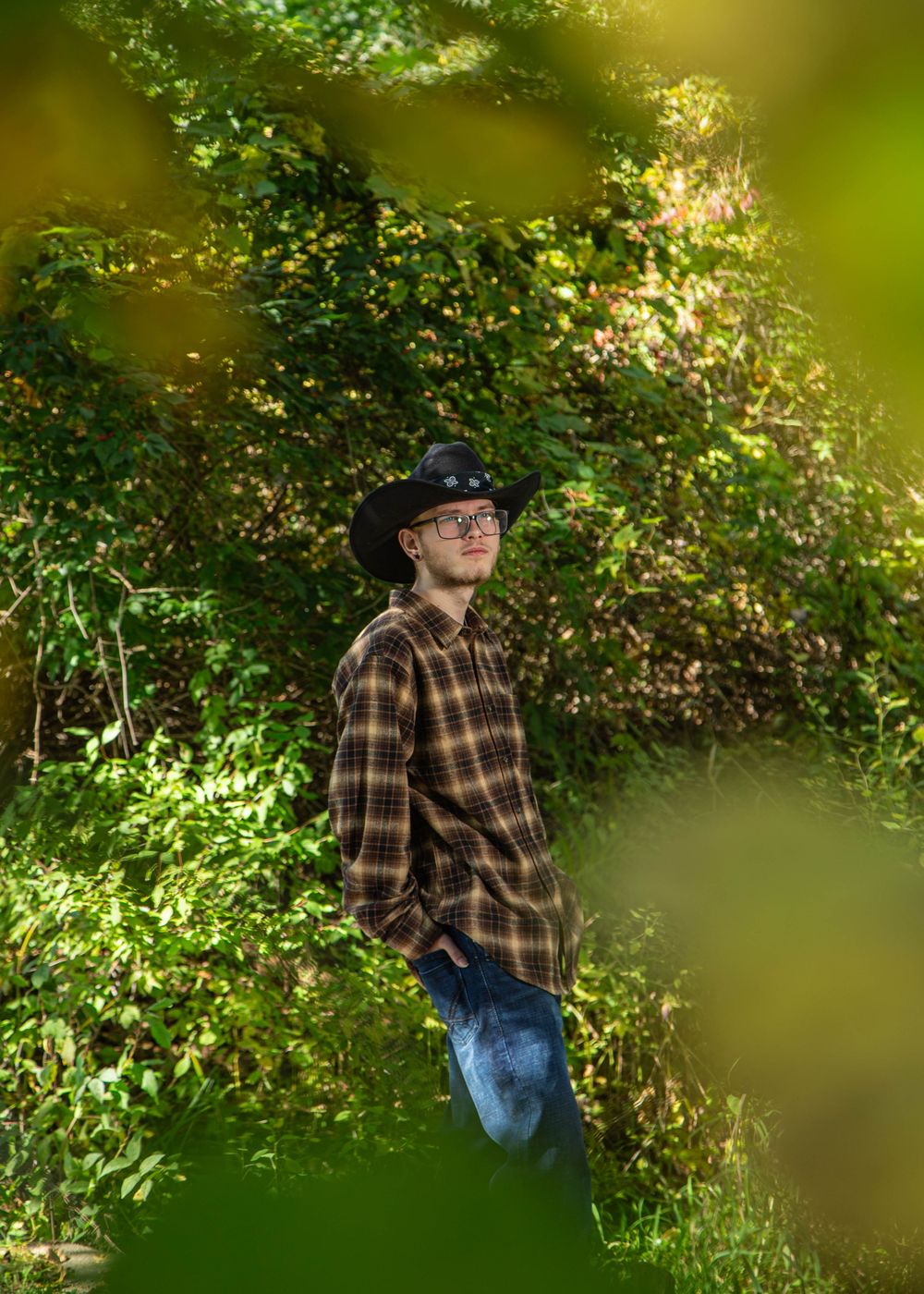 Young man in plaid shirt and black cowboy hat standing outdoors among lush green foliage in natural light.