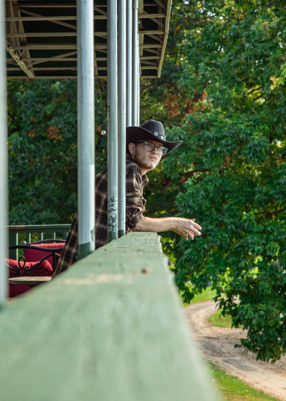 Man in cowboy hat and plaid shirt leaning on green railing of elevated structure with trees in background.