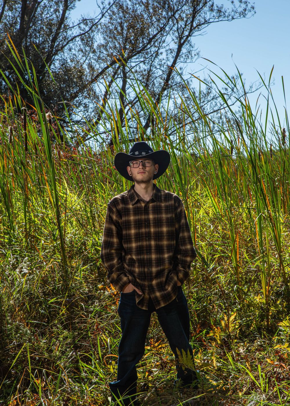 Young man in black cowboy hat and plaid shirt standing in tall green grass outdoors.