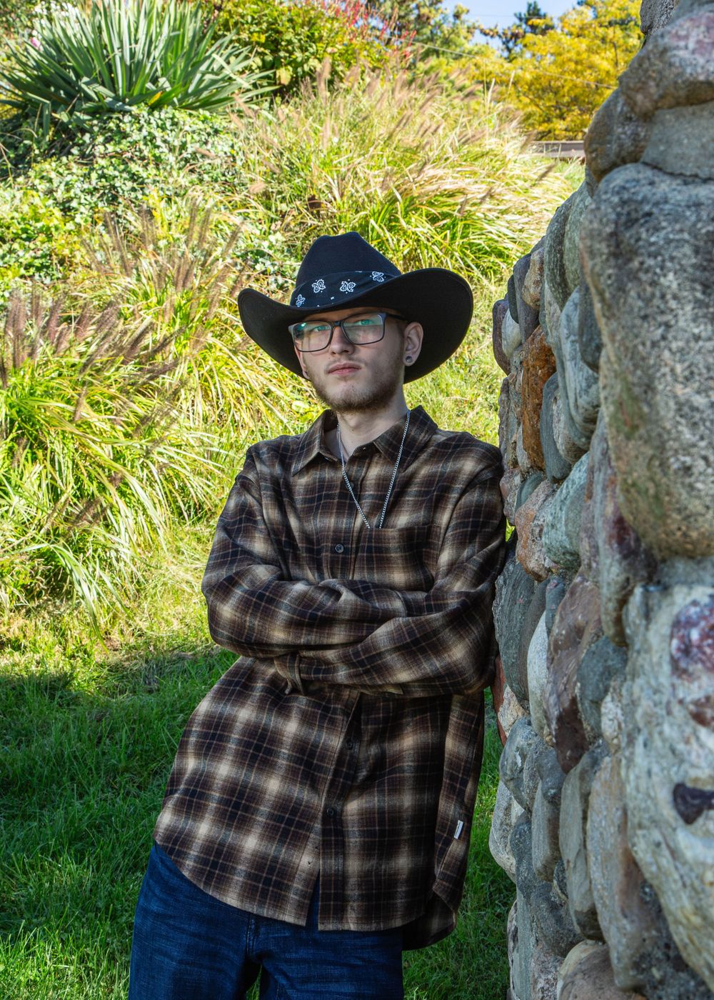 Young man in black cowboy hat and plaid shirt stands arms crossed beside stone wall in lush garden.