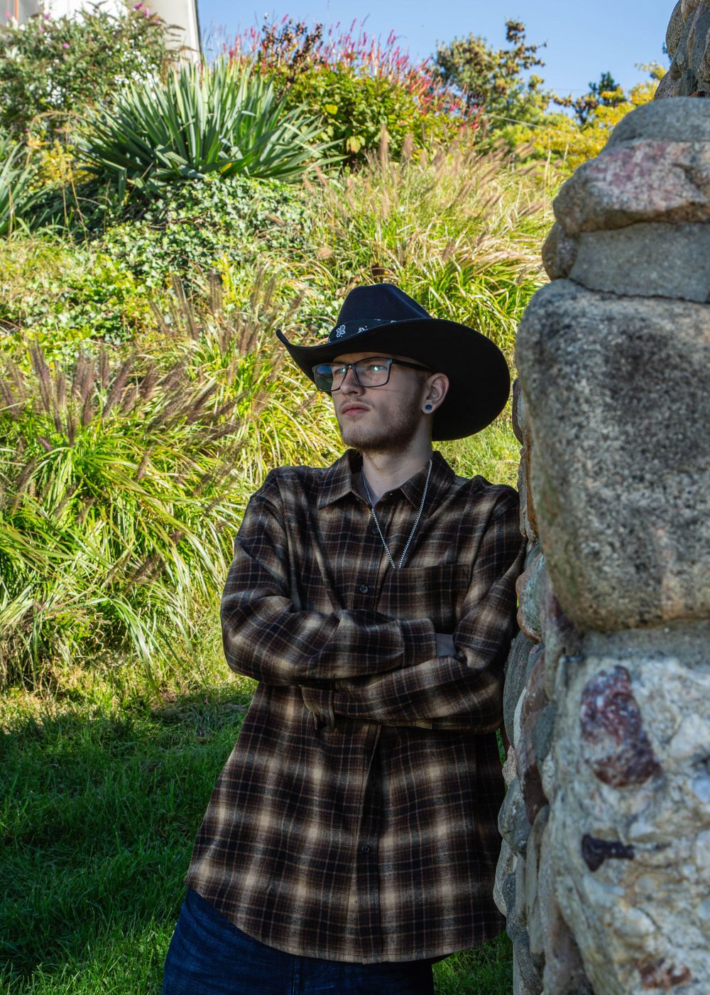 Young person in black cowboy hat and plaid flannel shirt leaning against stone wall in lush garden.