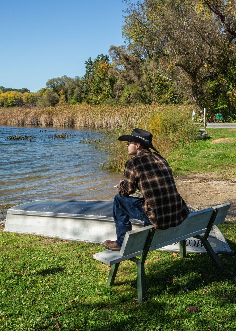 Person in cowboy hat and plaid shirt sits on bench gazing at calm lake surrounded by autumn trees on sunny day.