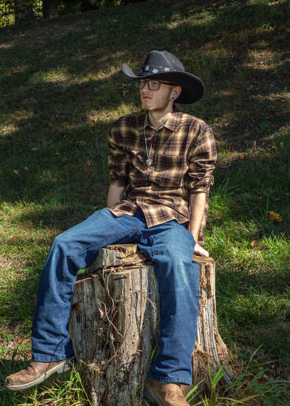 Young man in cowboy hat, plaid shirt, jeans, and boots sitting on a tree stump outdoors in sunlight.
