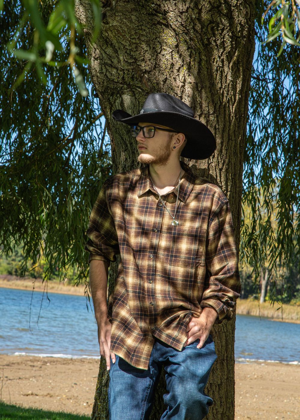 Young man in cowboy hat and plaid shirt leaning against a tree near a lake on a sunny day.