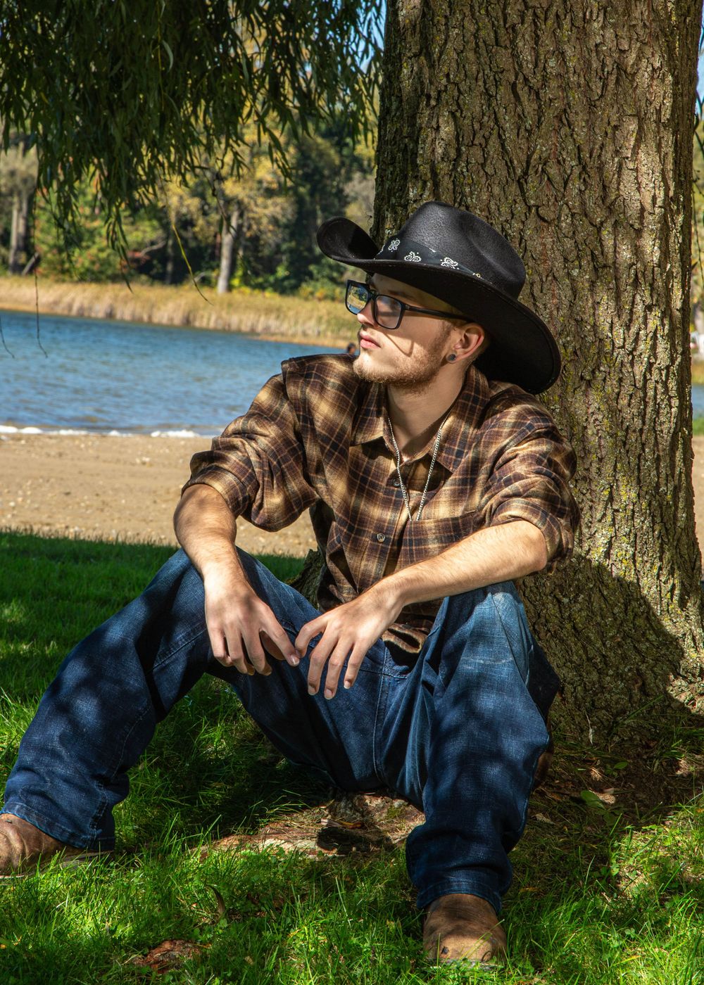 Young man in cowboy hat and plaid shirt sitting against tree by lakeside on sunny day.