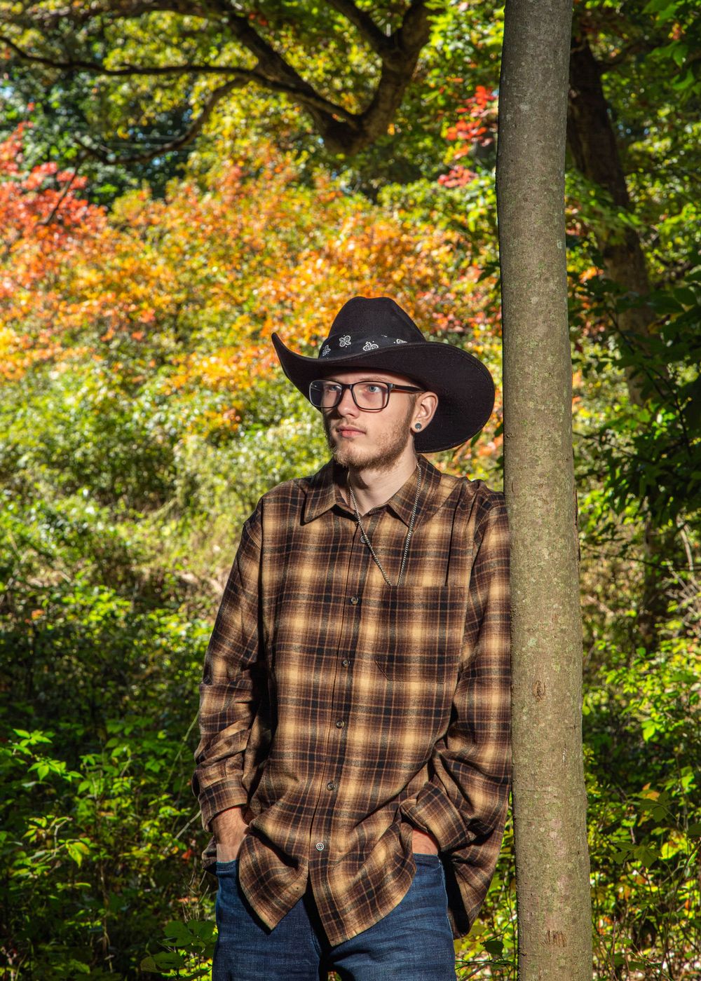 Young man in cowboy hat and plaid shirt poses outdoors among colorful autumn trees in fall foliage setting.
