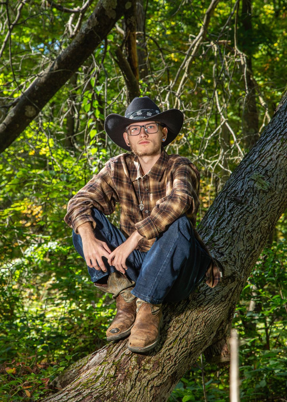 Young man in cowboy hat, plaid shirt, jeans, and boots sits on a tree in a lush green forest setting.