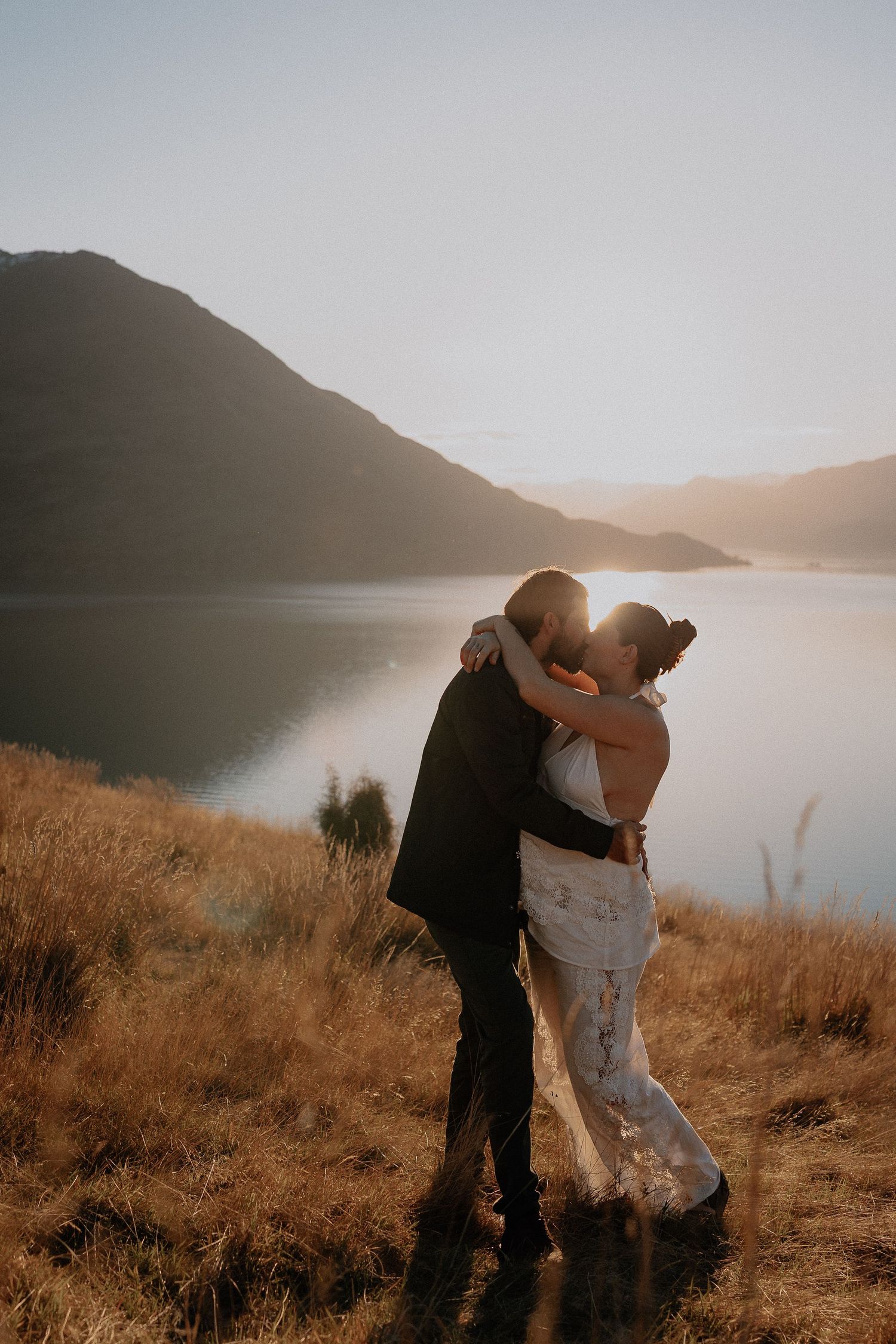 Couple sharing a romantic kiss at golden hour beside a serene mountain lake, bride in lace gown.